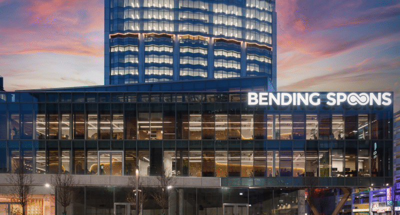 Modern glass office building at dusk with "BENDING SPOONS" illuminated on the facade. A taller building with distinctive lighting is visible behind it, and the sky shows hues of pink and blue at sunset.