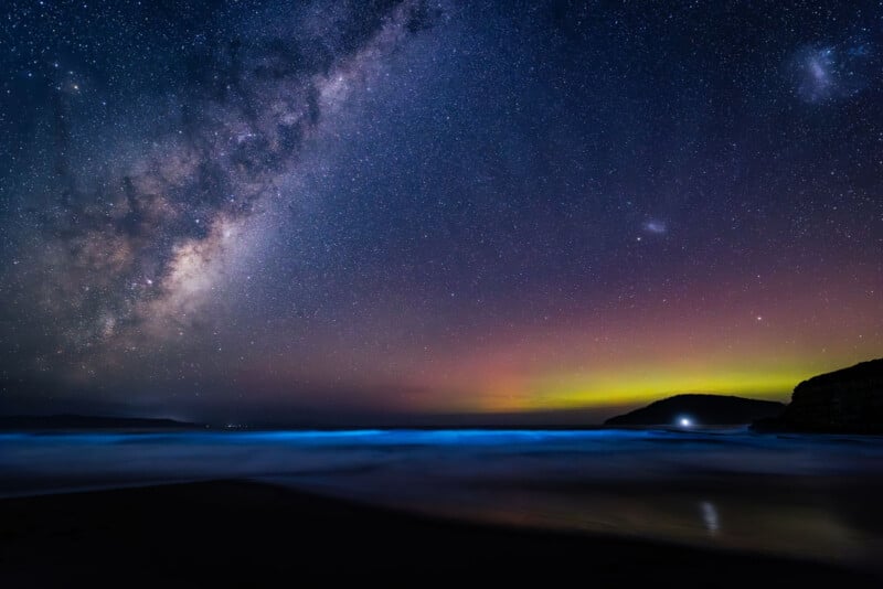 A vibrant night sky filled with stars and the Milky Way stretches above a calm ocean shore, with glowing bioluminescent waves and a faint yellow-green aurora near the horizon. Dark cliffs frame the scene.