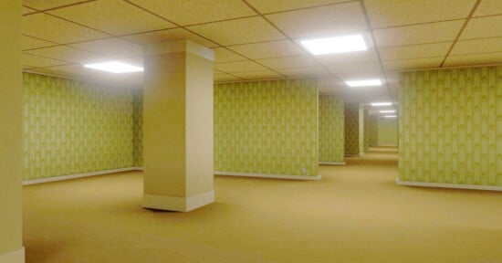 A large, empty room with yellowish carpet, beige ceiling tiles with bright fluorescent lights, and pale green patterned wallpaper. Several square columns are spaced throughout the open, maze-like space.