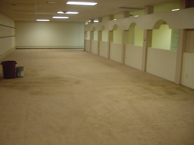 A large, empty room with beige carpet, white walls, and a row of archways along one side. Two trash bins and a bucket are positioned near the far end. The room is brightly lit by ceiling lights.