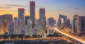 Cityscape at dusk with tall modern skyscrapers, brightly lit roads, and a distinctive building with an angular design. The sky is a gradient of orange and blue, indicating sunset.