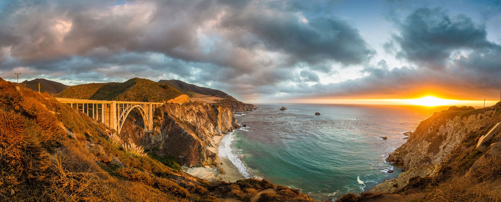 Panoramic view of Bixby Creek Bridge on California’s Big Sur coast at sunset, with rugged cliffs, green hills, and the Pacific Ocean beneath a dramatic sky.