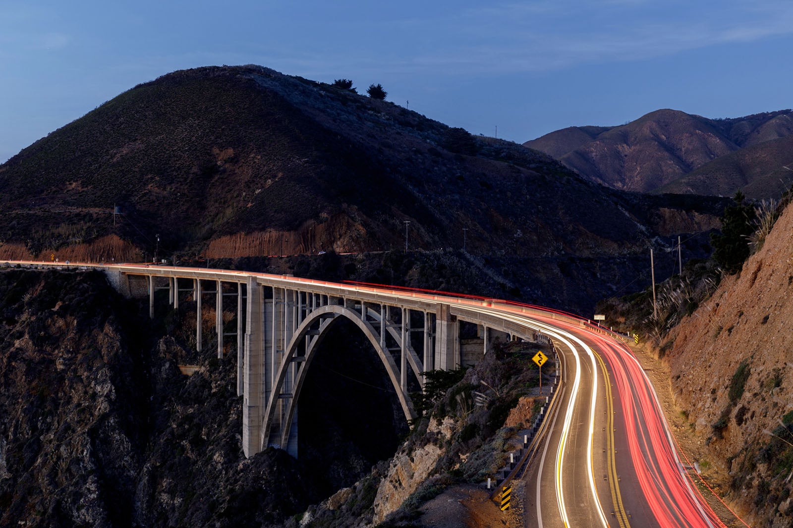 A curving bridge spans a deep canyon at dusk, with car headlights and taillights creating light trails along the road. Rugged, dark hills rise in the background under a dim sky.