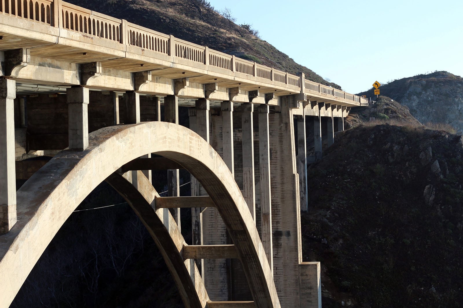 A concrete arch bridge spans a deep, rocky canyon with hillsides covered in sparse vegetation under a clear sky. The bridge features vertical supports and a curved arch beneath the roadway.