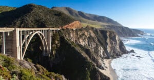 A tall arched bridge spans a rugged cliffside along a scenic coastline, with mountains and greenery in the background and waves crashing on a sandy beach below.
