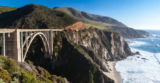 A tall arched bridge spans a rugged cliffside along a scenic coastline, with mountains and greenery in the background and waves crashing on a sandy beach below.