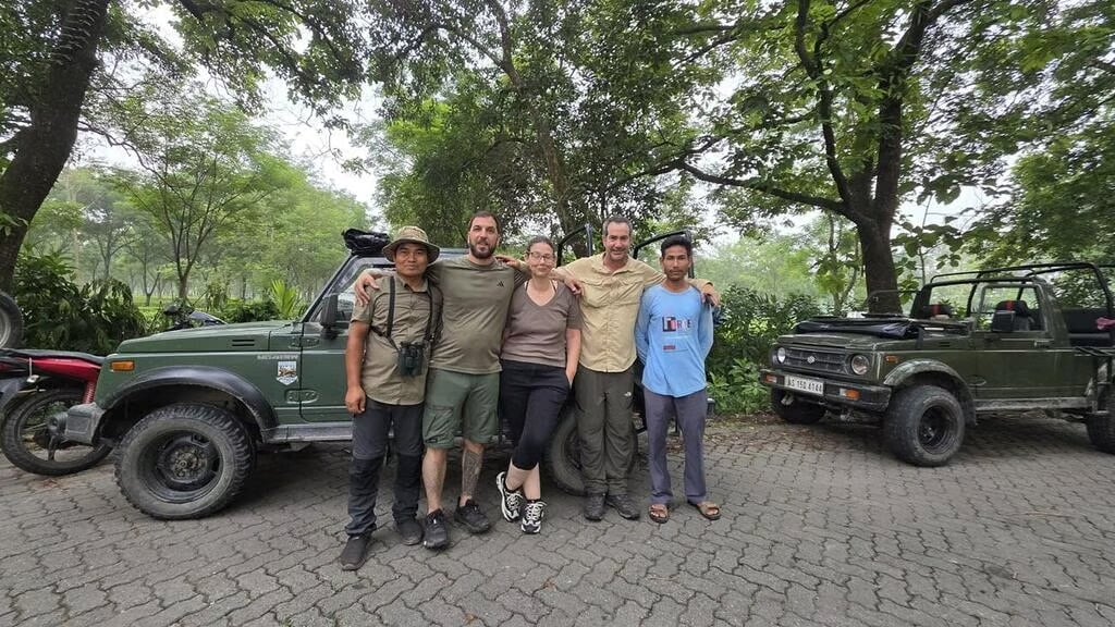 Five people stand smiling with arms around each other in front of two green jeeps in a lush, wooded area. They are dressed in outdoor or safari attire, suggesting they are on an expedition or tour.