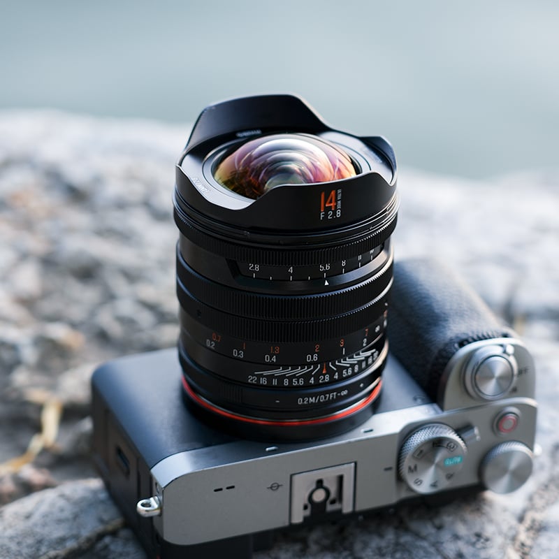 A close-up of a digital camera with a wide-angle lens, resting on a stone surface outdoors. The camera's dials and textured grip are visible, and the lens reflects colorful light.