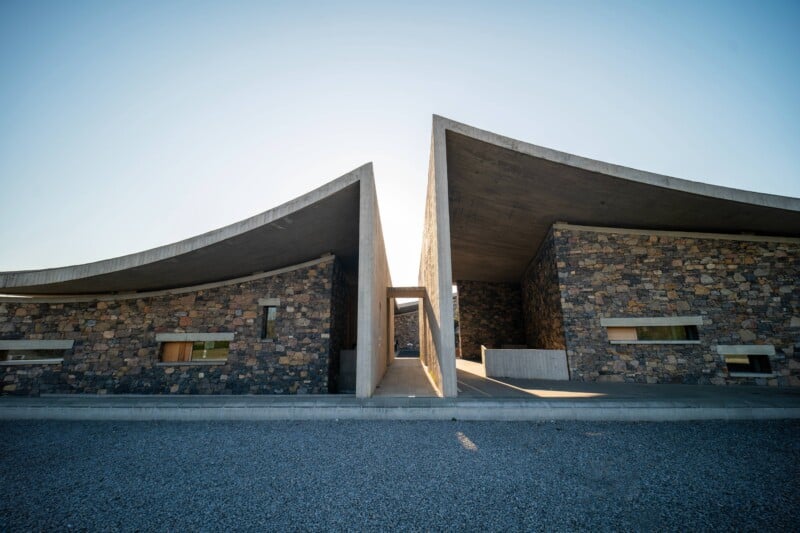 Modern building with two curved concrete roofs and stone walls, separated by a central walkway leading through the structure. The design creates a dramatic, symmetrical appearance under a clear sky.