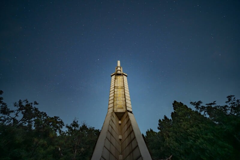 A tall, illuminated monument rises into a starry night sky, framed by dark trees on either side. The structure is viewed from a low angle, emphasizing its height and prominence.