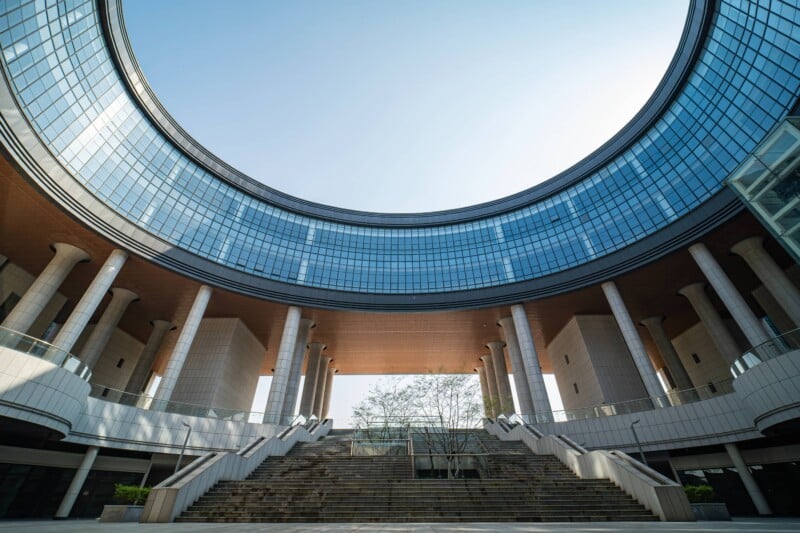 Modern building with a large circular glass facade, tall columns, and wide stone steps leading to an open courtyard, viewed from below on a clear day.