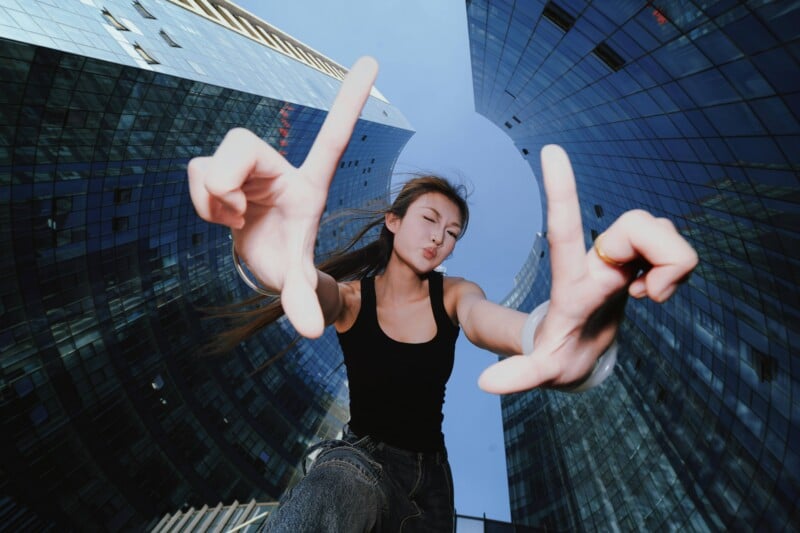 A young woman in a black tank top and jeans makes a "peace" sign with her fingers, pouting her lips, against a backdrop of modern glass skyscrapers.