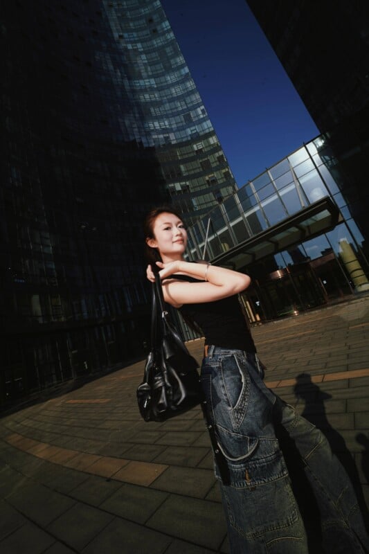 A woman in a black tank top and wide-leg jeans stands outdoors, holding a black shoulder bag, with tall glass buildings and clear blue sky in the background.