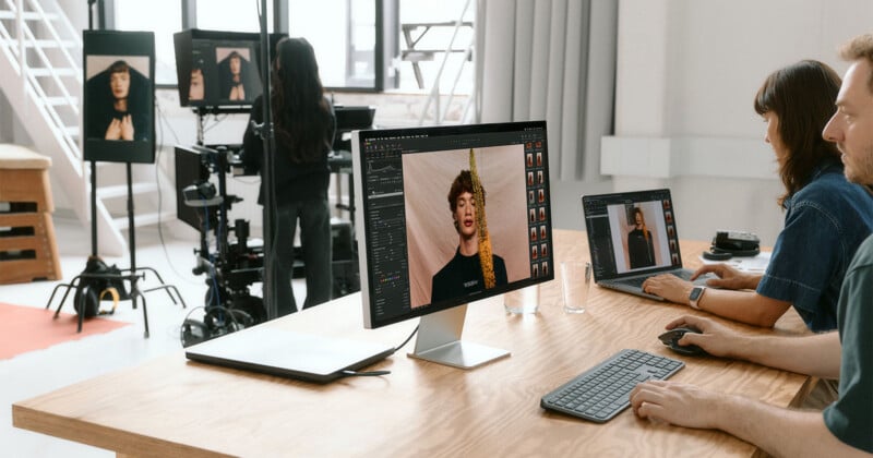 A modern photo studio with two people editing portraits on computers at a wooden desk, while another person photographs a model posing in front of a camera and lighting setup in the background.