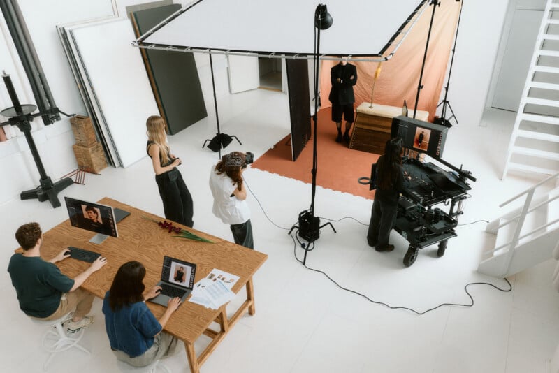 A photography studio scene with a model posing on an orange backdrop, while a team operates cameras and computers, and others observe or work on laptops at a table. Bright lighting equipment surrounds the set.