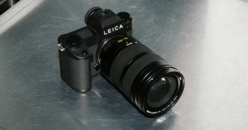 A black Leica digital camera with a large zoom lens rests on a metallic surface, viewed at an angle that shows the camera body and lens details clearly.