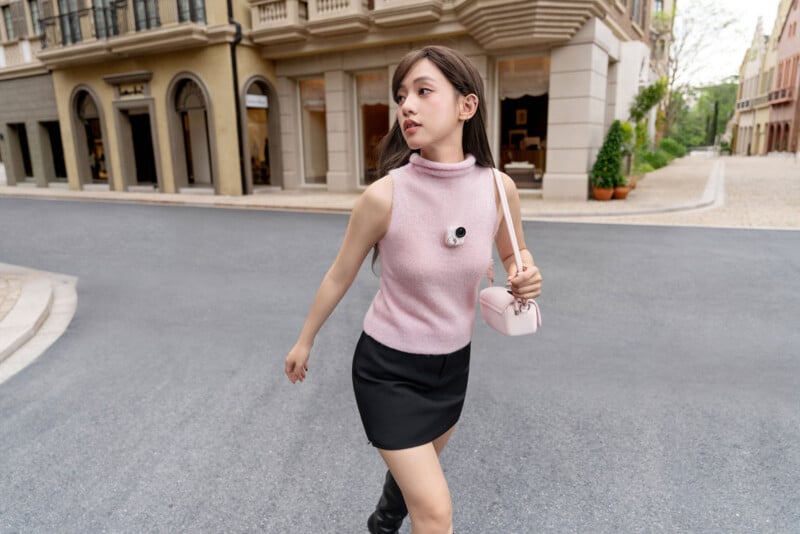 A young woman wearing a sleeveless pink turtleneck top, black skirt, and carrying a small pink purse walks across a quiet street in an urban area with stylish buildings in the background.