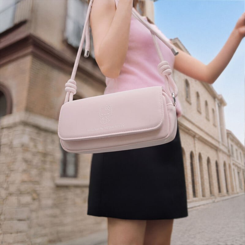 A person wearing a pink sleeveless top and black skirt holds a light pink Hello Kitty shoulder bag while standing on a street with stone buildings in the background.