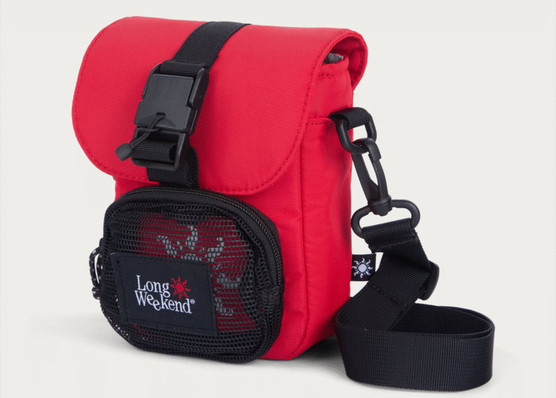 A small red crossbody bag with a black adjustable strap, black buckle closure, and a front mesh pocket labeled "Long Weekend" with a red sun graphic. The bag has a modern, functional design.