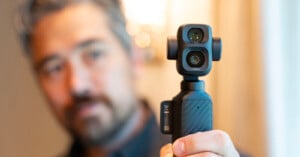 A man with gray hair and a beard holds a small, handheld camera with dual lenses, focusing on the device while the background is blurred.