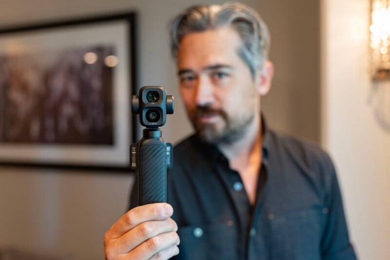 A man with gray-streaked hair and a beard holds a small handheld camera towards the viewer, with the background softly out of focus.