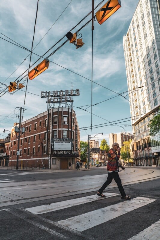 A man in a plaid shirt crosses a city street at a crosswalk near a brick building with a large “FILMORE’S HOTEL” sign. Tall modern buildings and overhead wires are visible under a blue sky.