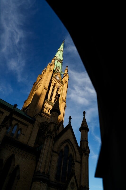 A tall, pointed church steeple with a green spire rises against a blue sky with wispy clouds, partially framed by a dark shadow in the foreground. Warm sunlight illuminates the building's stone facade.
