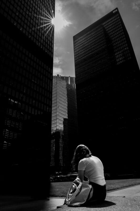 A woman sits alone with a backpack in a city plaza, surrounded by tall glass skyscrapers. Sunlight shines through the clouds, casting dramatic shadows across the scene.