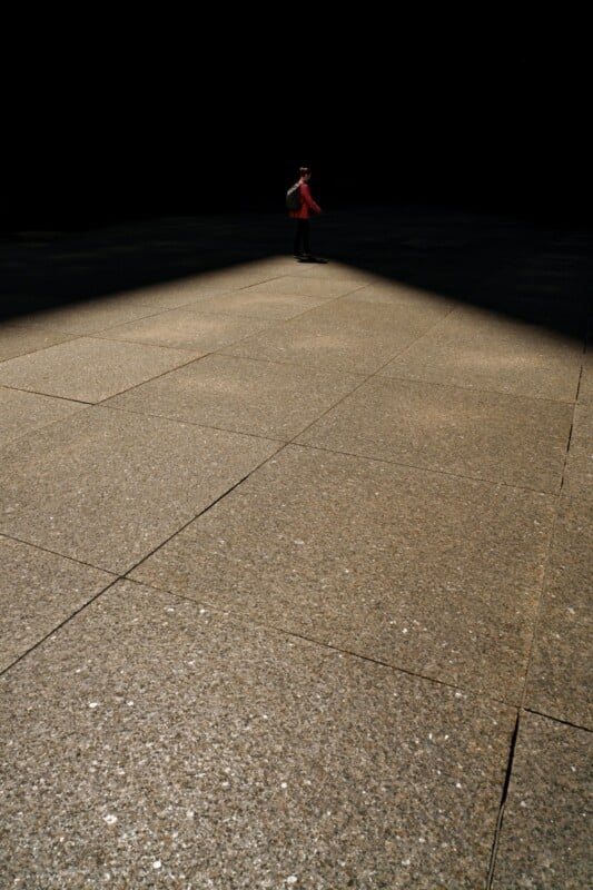 A person wearing a red jacket and backpack stands alone in a beam of light on a large, empty, textured concrete surface with darkness surrounding them.