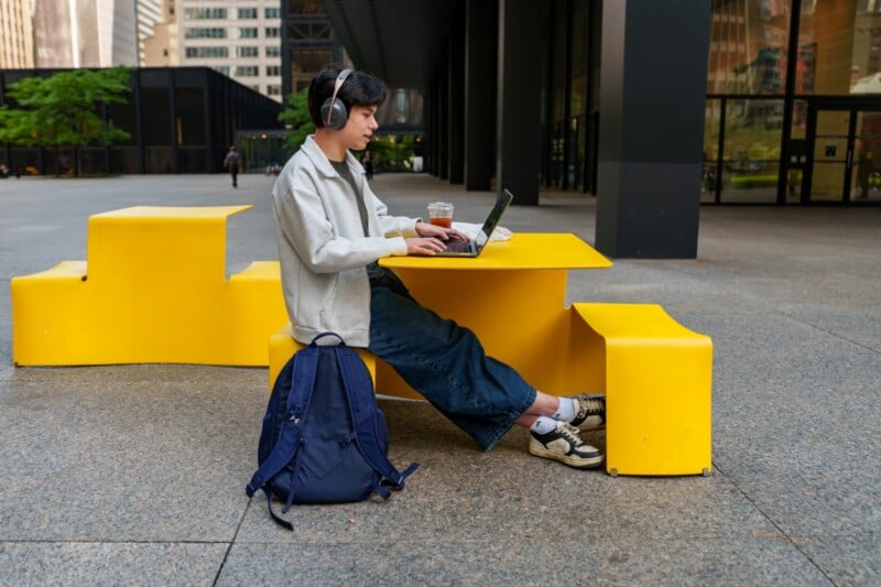 A person wearing headphones works on a laptop at a bright yellow outdoor table in an urban setting. A coffee cup and backpack are nearby, and tall buildings are visible in the background.