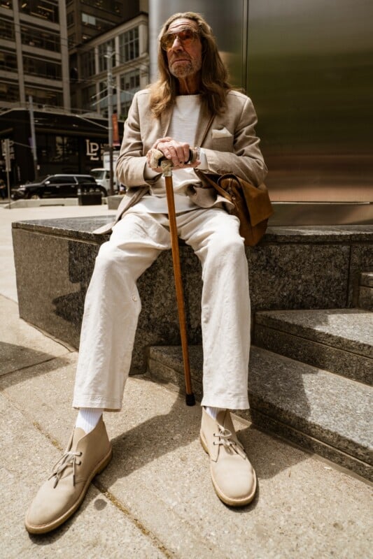 An older man with long hair, wearing sunglasses, a beige blazer, light pants, and cream shoes, sits on stone steps outdoors holding a wooden cane, with city buildings in the background.