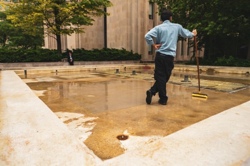 A person wearing dark pants and a light blue shirt uses a broom to clean an empty concrete fountain or pool in an outdoor urban area, with greenery and buildings in the background.