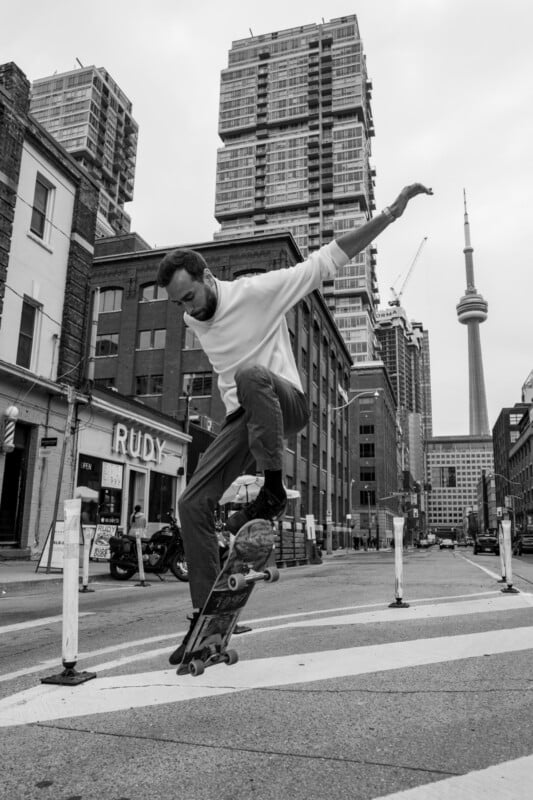 A man in mid-air performs a skateboard trick on a city street, surrounded by tall buildings. The CN Tower is visible in the background. The scene is black and white, with a lively urban atmosphere.