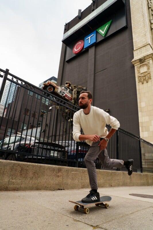 A man in a white sweater and gray pants rides a skateboard on a city sidewalk. Behind him is a black fence, a decorative car sculpture, and a tall building with a large CTV sign.