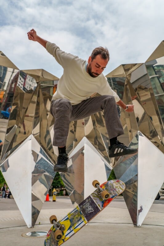 A man in a white sweater and gray pants performs a trick on a skateboard in front of a geometric metallic sculpture in an urban setting.