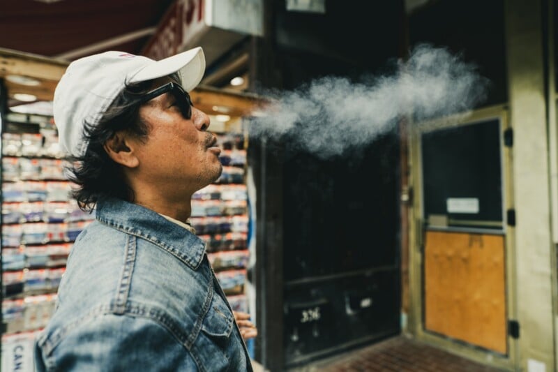 A person wearing a white cap, sunglasses, and a denim jacket exhales a cloud of smoke while standing on a city sidewalk in front of a store with a glass door and display shelves.