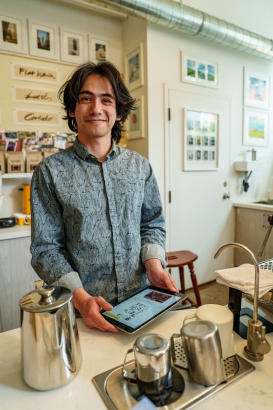 A smiling barista with dark hair stands behind a coffee counter, holding a tablet. Coffee equipment, cups, and framed pictures decorate the cozy cafe setting.