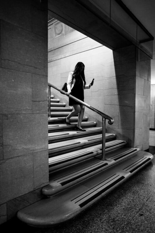 A woman in a dress walks down a staircase while looking at her phone in a dimly lit, modern building. The scene is in black and white.