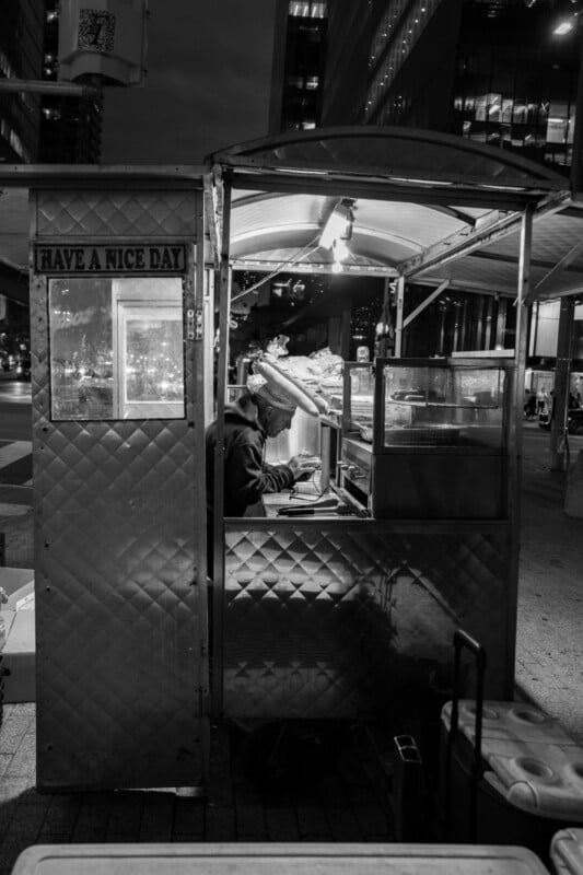 A black-and-white photo of a person working inside a small street food cart at night, surrounded by city lights and tall buildings, with a sign on the cart that reads "Have a Nice Day.