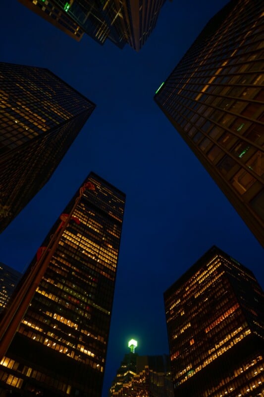 Nighttime view looking up at tall office buildings with illuminated windows, forming a circle against a dark blue sky; a single lit dome or spire is visible in the distance at the bottom center.