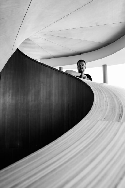 A man stands behind a curved wooden railing in a modern building with sweeping architectural lines, illuminated by natural light. The photo is in black and white.