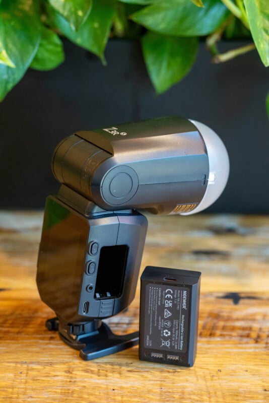 A black external camera flash unit standing on a wooden surface with its battery compartment open. The rechargeable battery is placed beside it. Green leaves are visible in the blurred background.