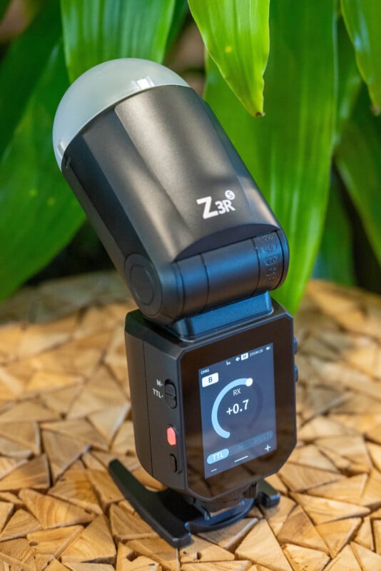 A black Z3R camera flash with a white diffuser is positioned on a wooden surface, with green leaves in the background. The digital screen on the flash displays various settings and information.