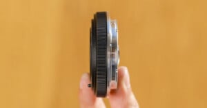 A hand holds a thin camera lens adapter vertically against a blurred, warm-toned background.
