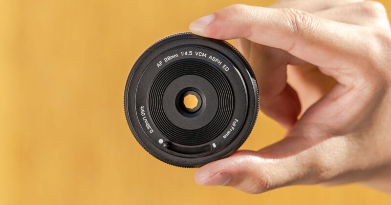 A hand holding a camera lens with text around the front rim reading "AF 28mm 1:4.5 VCM ASPH ED" against a blurred yellow background.