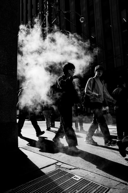 People walking on a city street as steam rises from a grate, with dramatic light and shadows; most of the figures are in silhouette, and tall buildings are visible in the background.