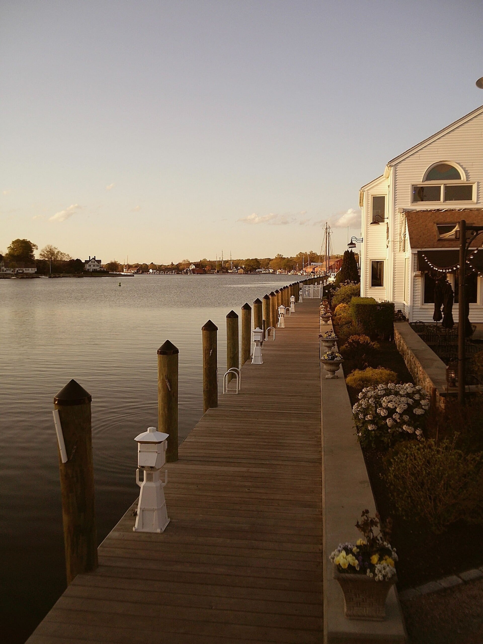 A wooden dock with white lanterns runs alongside a waterfront house, bordered by flowers and shrubs, with calm water and boats visible in the background under a clear sky at sunset.