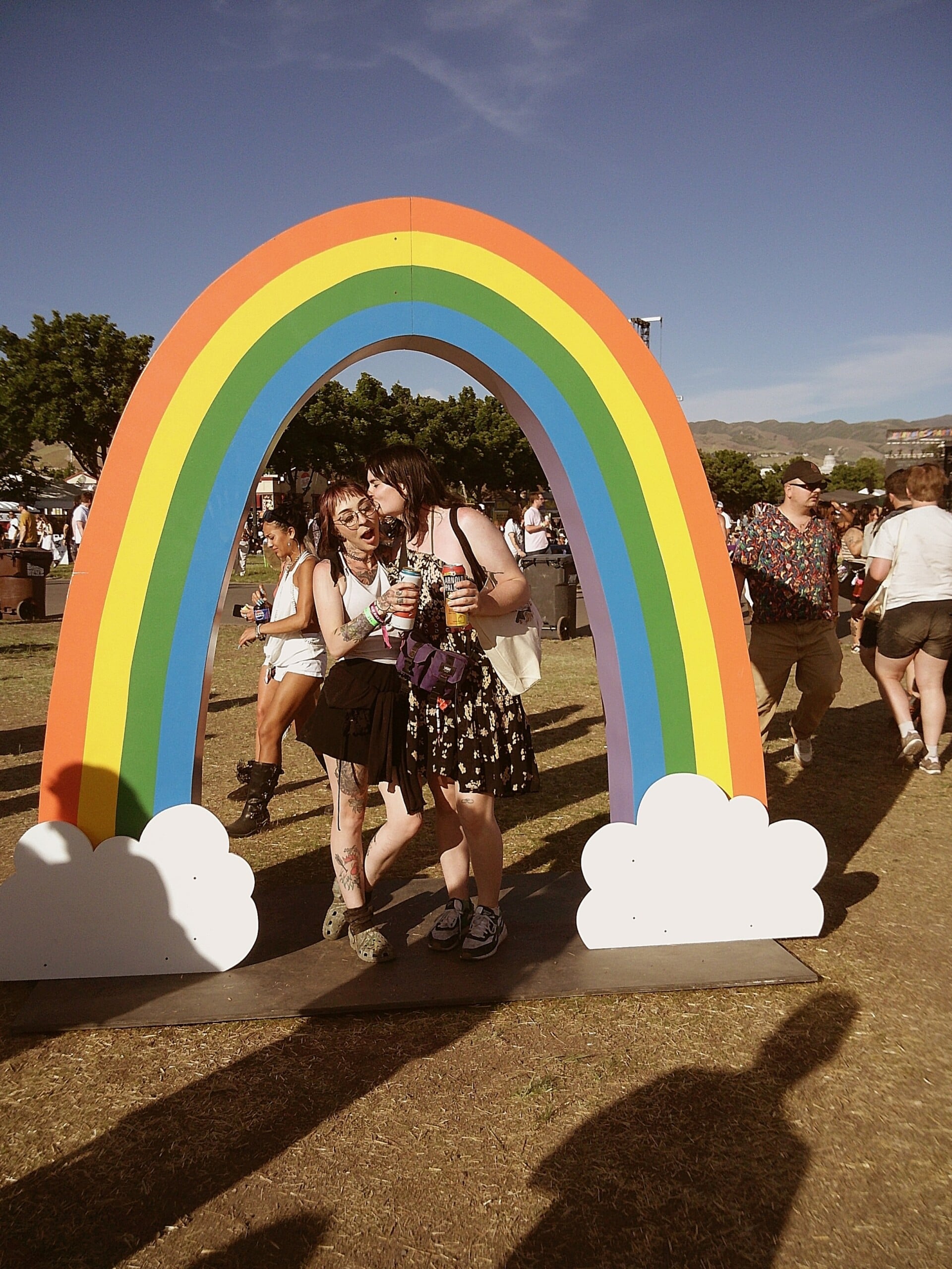 Two people pose together under a colorful rainbow arch with clouds at an outdoor festival. Other festival-goers and trees are visible in the background under a clear blue sky.