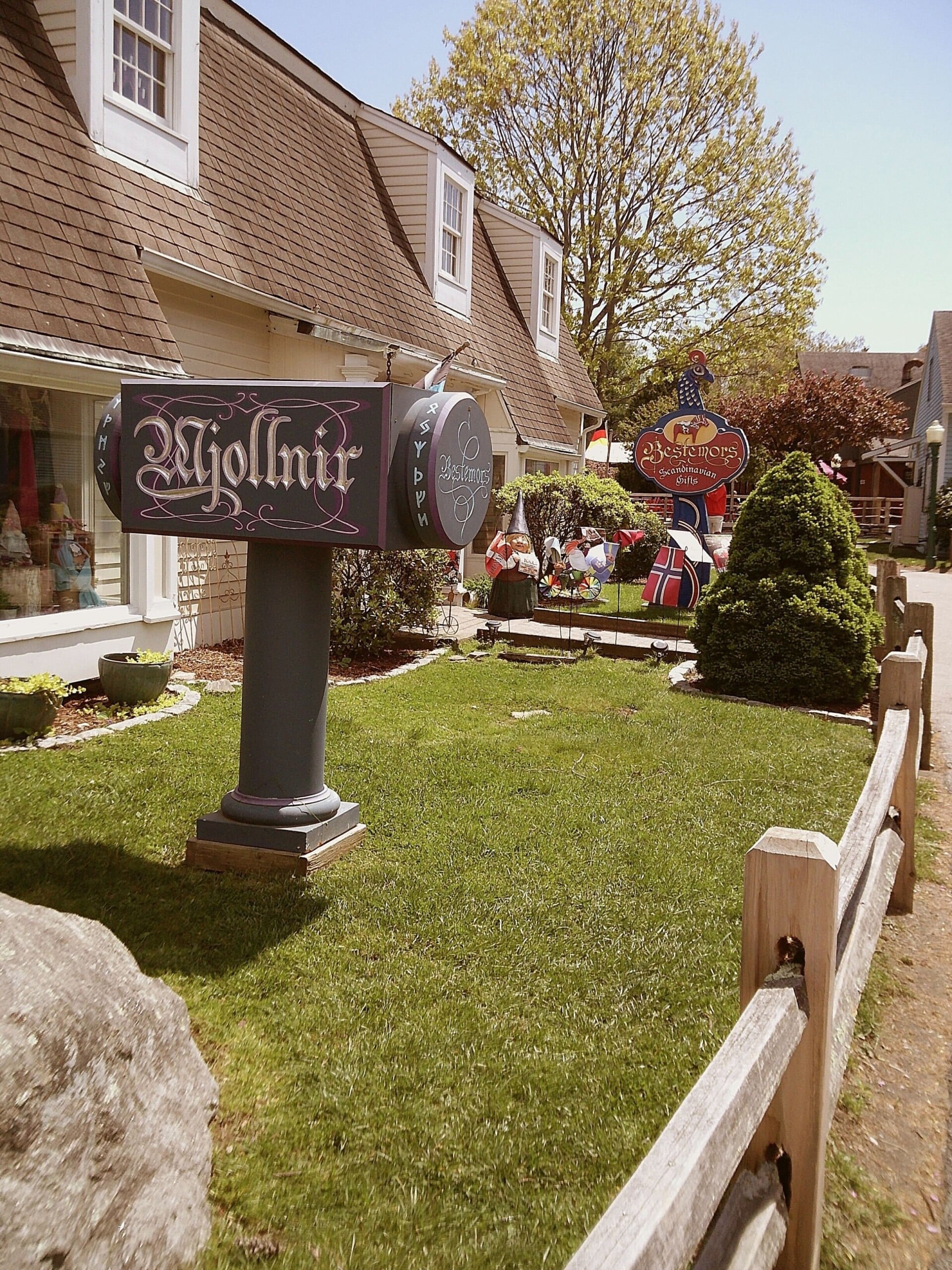 A mailbox labeled "Mjolnir" stands in front of a quaint building with Nordic-themed decorations, including flags and painted cutouts, on a neatly kept lawn bordered by a wooden fence.