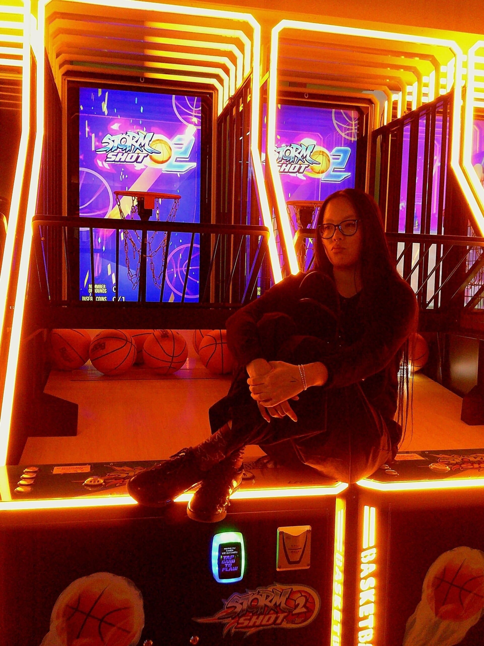 A person with long dark hair and glasses sits on the edge of a brightly lit arcade basketball game, surrounded by glowing orange lights and basketballs, with digital screens in the background.